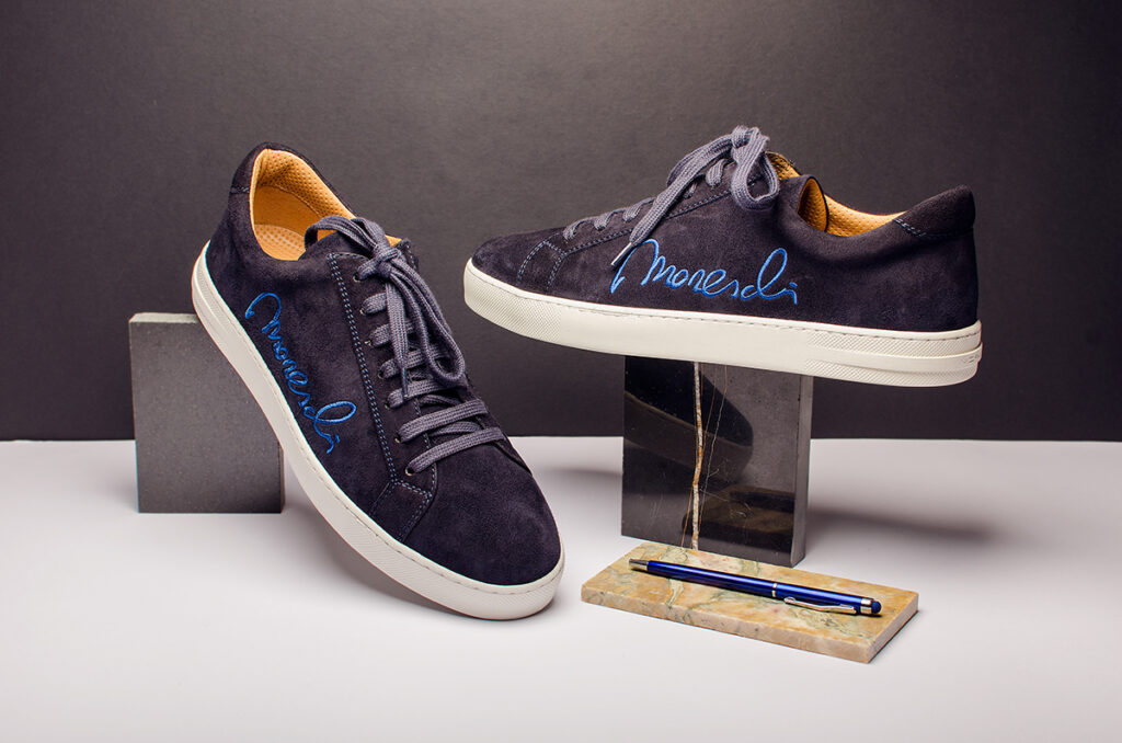 Fotografia still-life – scarpe da uomo Moreschi in camoscio blu scuro con ricamo blu elettrico, esposte su un piedistallo di marmo.