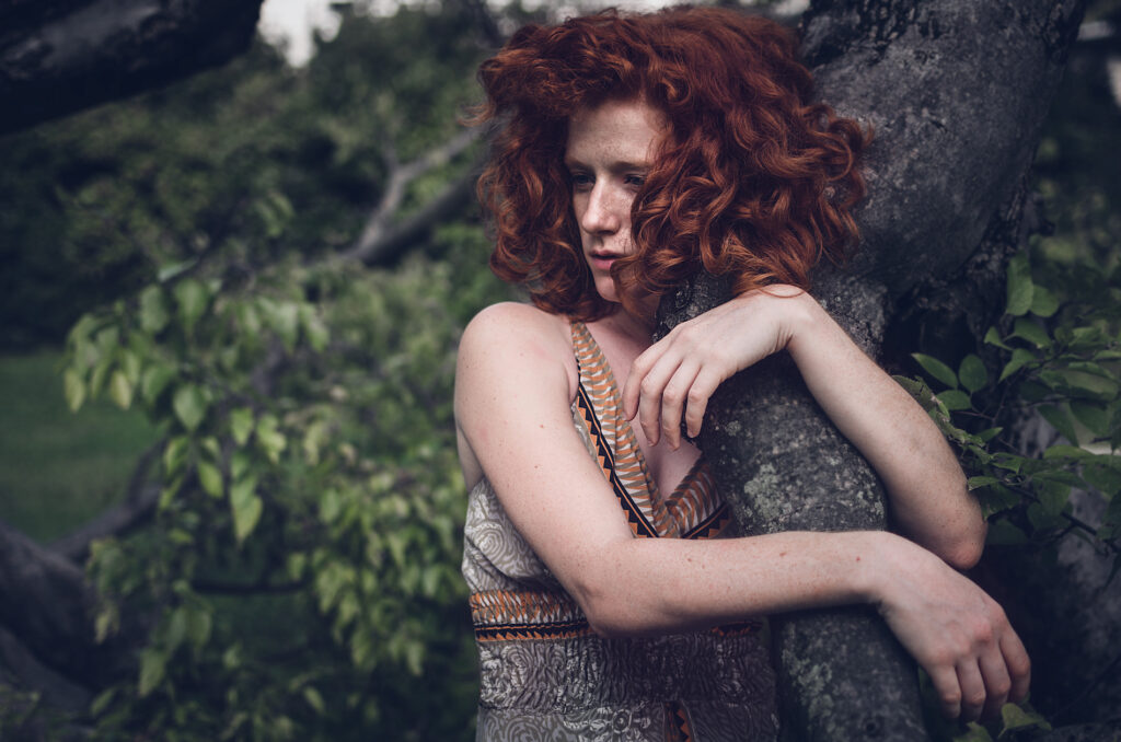 Modella dai capelli rossi avvolgenti posa abbracciata a un albero in un vestito patterned, immersa nella natura lussureggiante.