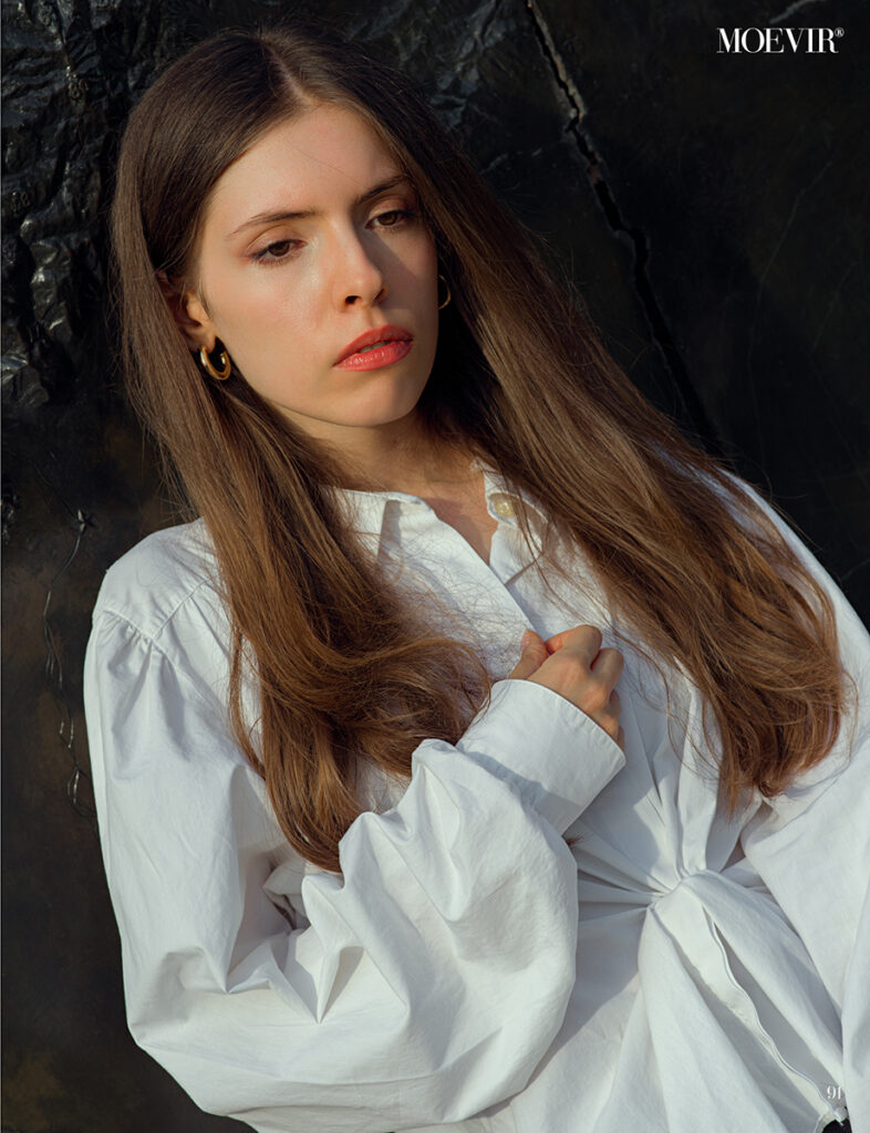 Modella con capelli castani lunghi indossa una camicia bianca oversize con dettagli eleganti, posa in una luce naturale morbida con sfondo scuro.