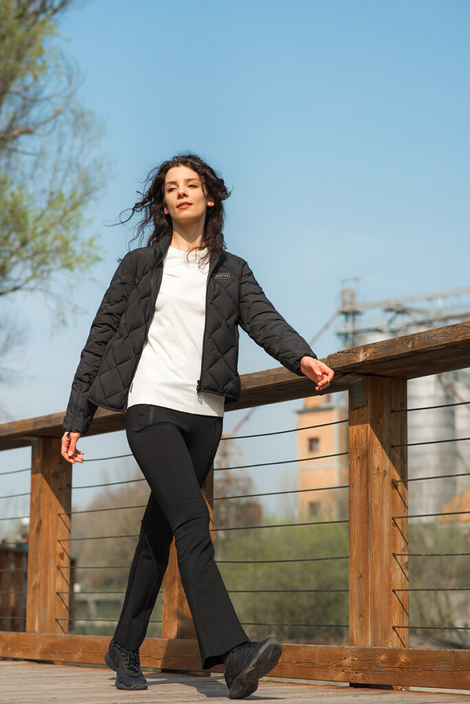 Modella in giacca imbottita nera e pantaloni sportivi Duvetica, posando su un ponte in legno in una giornata soleggiata.