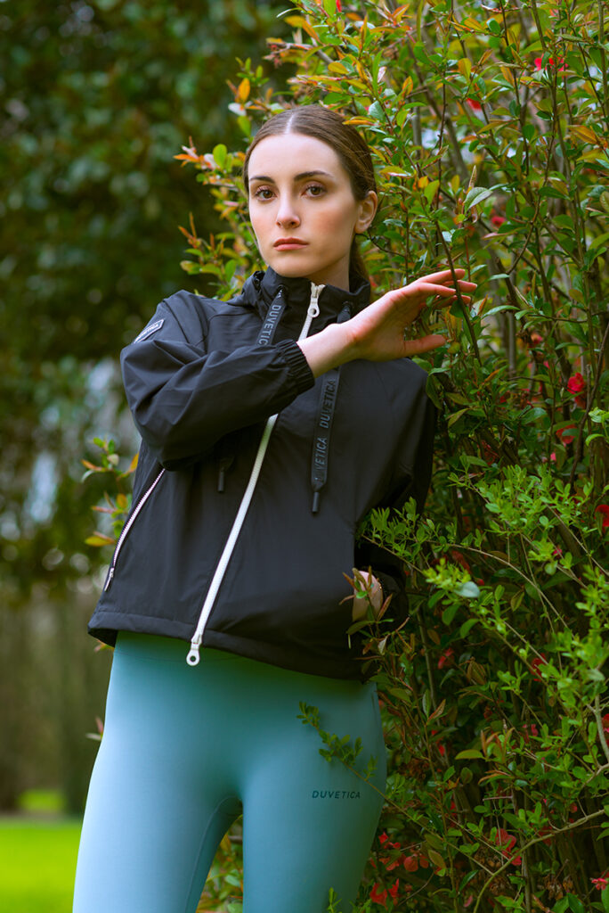 Modella in giacca sportiva nera e leggings azzurri Duvetica posa in un giardino fiorito, fotografia di Marius Cruceriu.