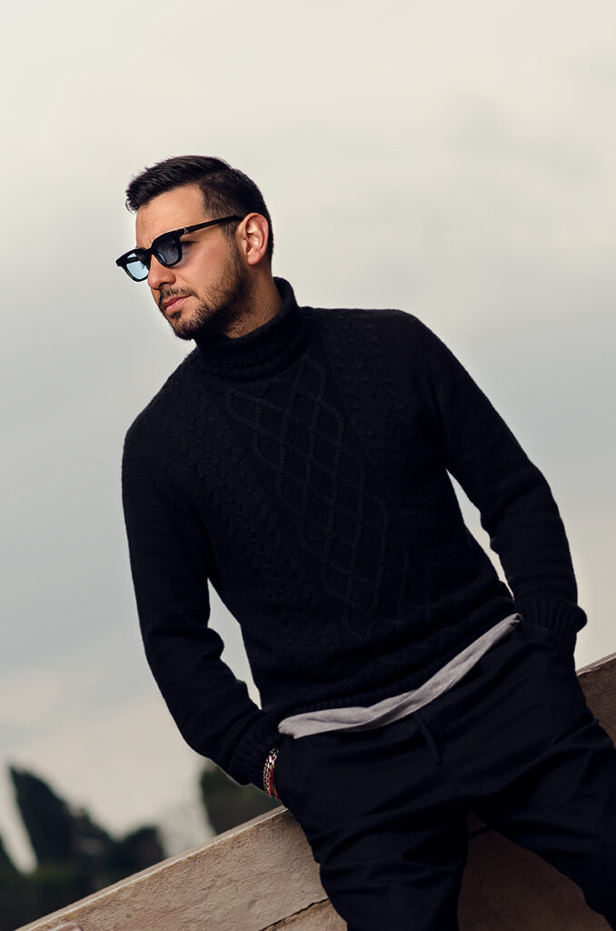 Uomo con occhiali da sole artigianali in posa casual su sfondo urbano, scatto lifestyle moda