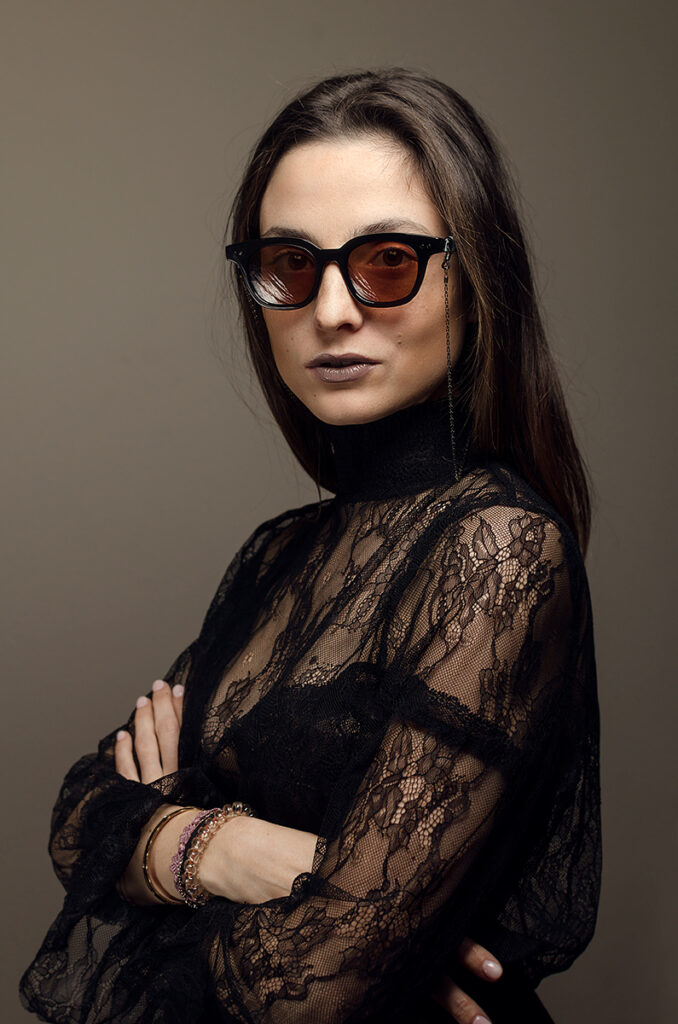 Ritratto editoriale di donna con occhiali da sole artigianali e top in pizzo nero, fotografia moda elegante