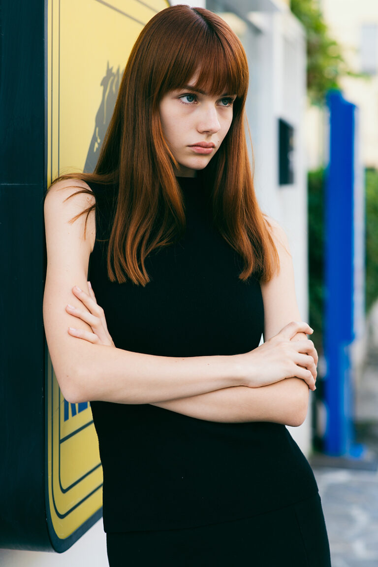 Ritratto a tre quarti della modella Agata durante un test per Tank Agency a Milano, in total black con top senza maniche e braccia conserte, appoggiata a un’insegna gialla in esterno, fotografata da Marius Cruceriu.