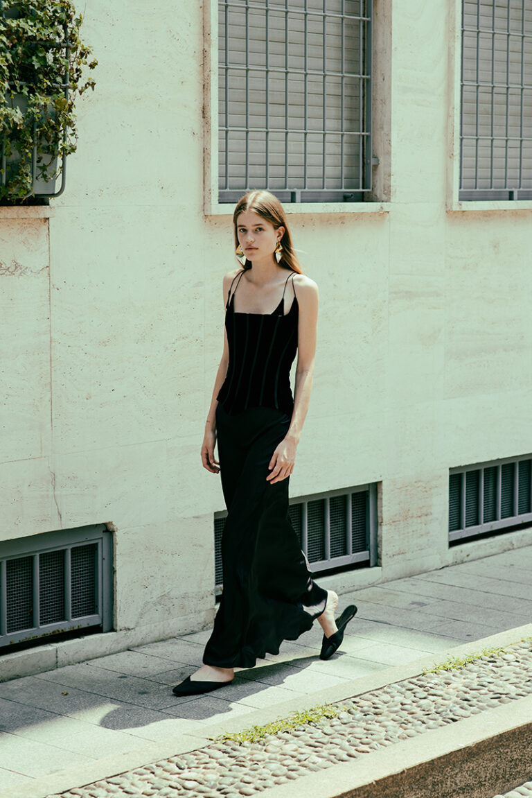 Modella Juta durante un test per Supreme Management Milano, ritratta mentre cammina lungo un marciapiede a Milano con abito nero lungo e top a spalline sottili, fotografata da Marius Cruceriu.