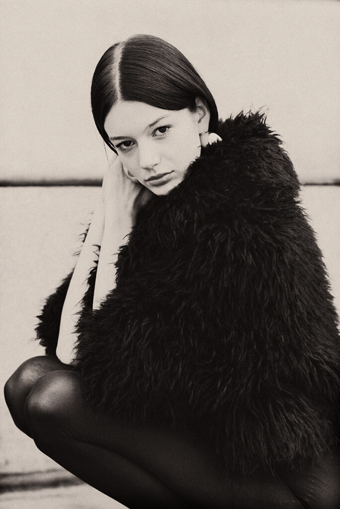 Modella Zuzanna durante un test per Supreme Management Milano, in posa accovacciata con cappotto nero in pelliccia e collant aderenti, fotografata da Marius Cruceriu.