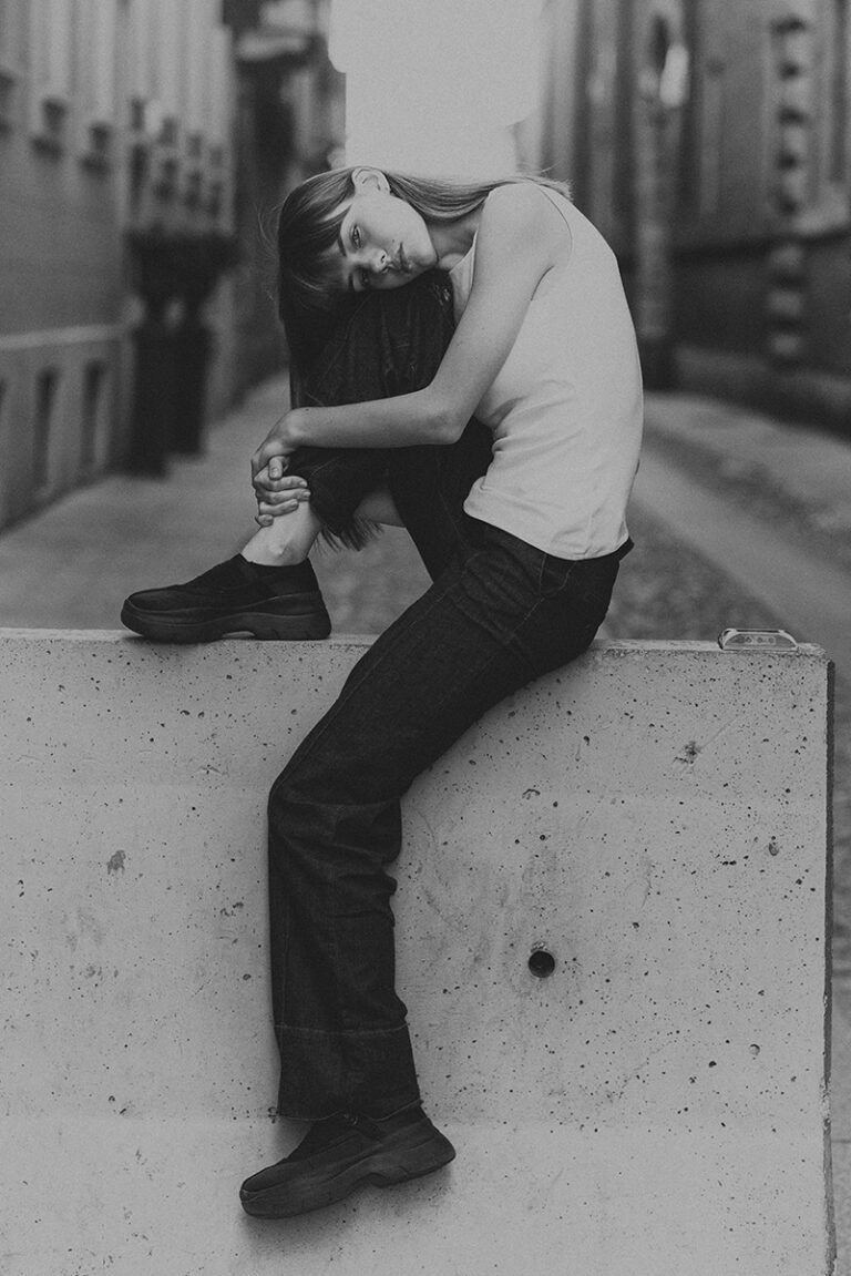 Modella Agata durante un test per Tank Agency a Milano, seduta su un blocco di cemento in una strada urbana, in bianco e nero con canotta bianca, jeans scuri e sneakers, fotografata da Marius Cruceriu.