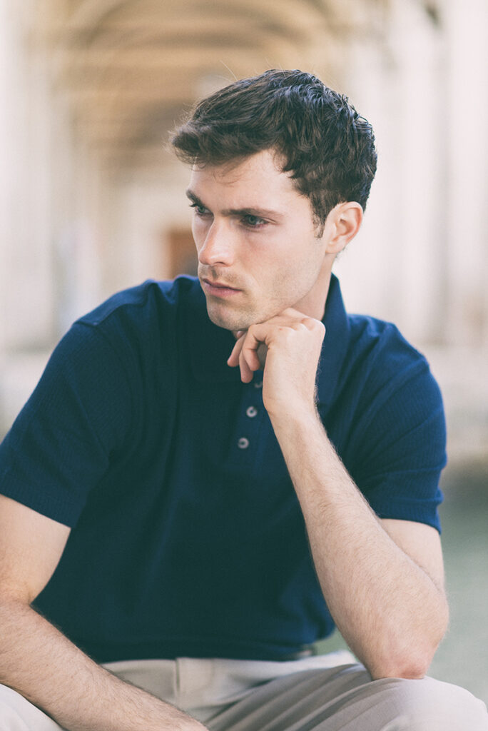Modello Blake seduto a Venezia con polo blu, in posa pensierosa con mano sotto il mento, ritratto maschile a colori di Marius Cruceriu.
