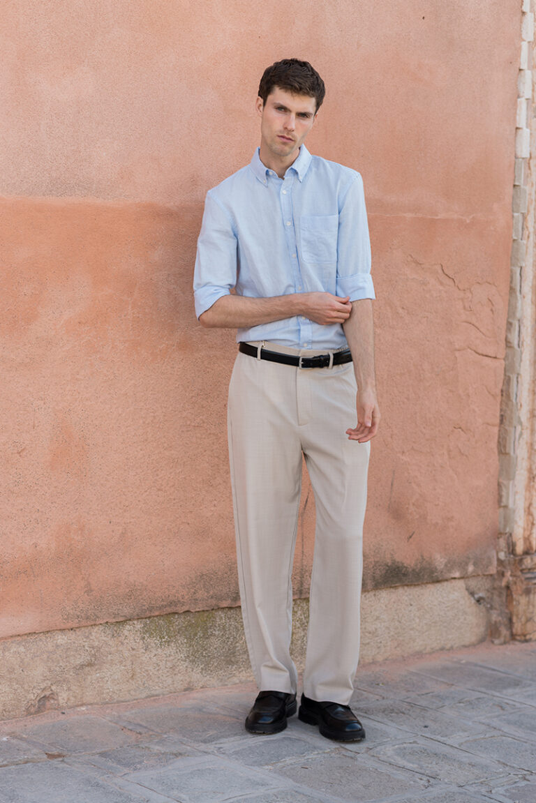 Modello Blake a Venezia in piedi a figura intera contro un muro color terracotta, con camicia azzurra e pantaloni chiari, fotografato da Marius Cruceriu.