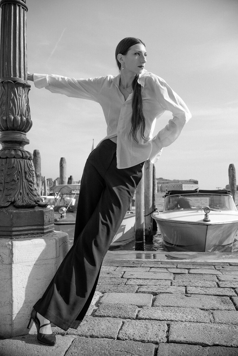 Modella Gloria a Venezia per Iso Model Management, scatto a figura intera in bianco e nero appoggiata a un lampione, look con camicia bianca e pantaloni neri, test model realizzato da Marius Cruceriu.