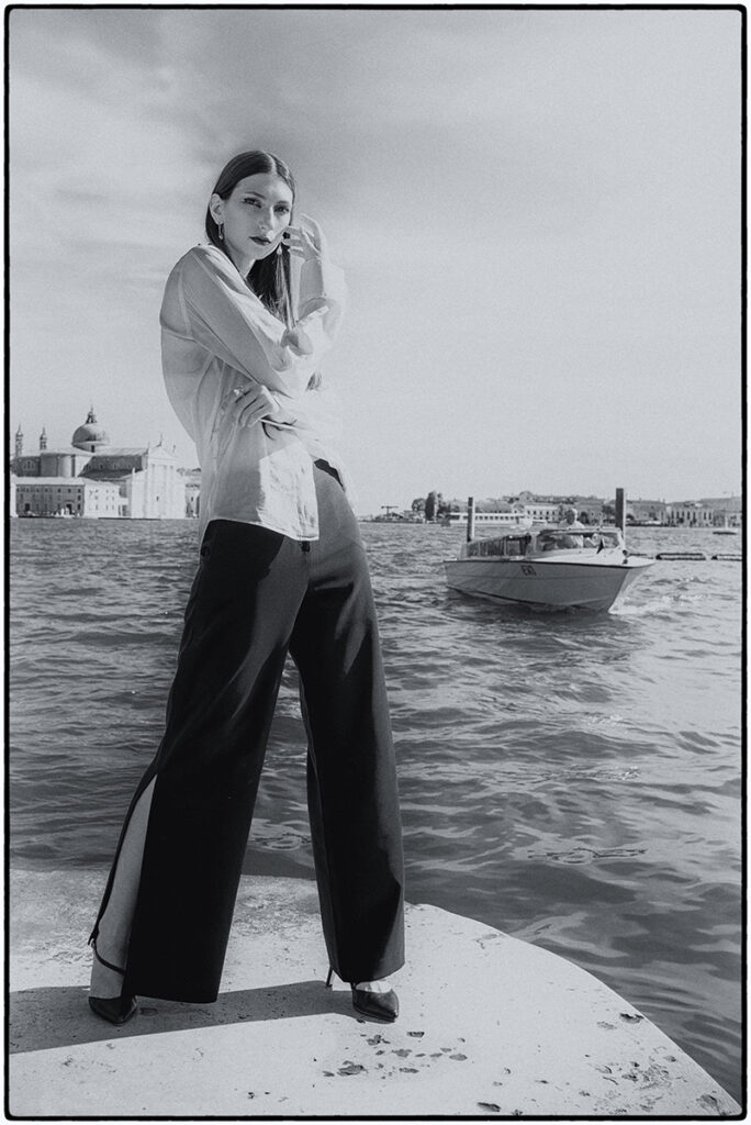 Modella Gloria a Venezia, scatto editoriale in bianco e nero sulla laguna con barca sullo sfondo, look con pantaloni ampi e camicia, fotografia e direzione artistica di Marius Cruceriu.