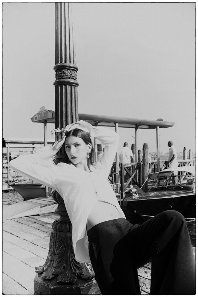 Modella Gloria a Venezia per Iso Model Management, scatto in bianco e nero street style, seduta su un molo con imbarcazioni sullo sfondo, indossa camicia bianca e pantaloni neri, test model realizzato da Marius Cruceriu.