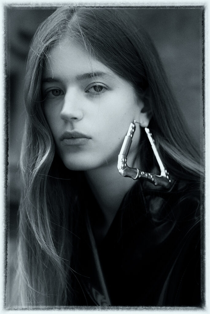 Primo piano in bianco e nero della modella Juta durante un test per Supreme Management Milano, con capelli lunghi sciolti, orecchino metallico scultoreo e giacca in pelle, fotografata da Marius Cruceriu.
