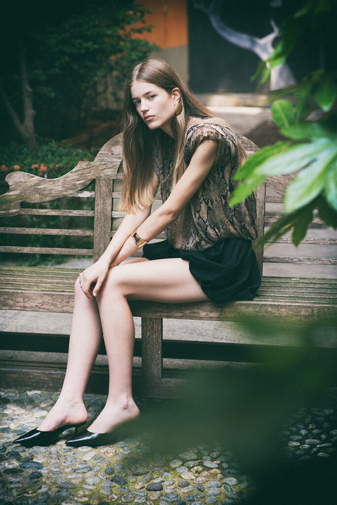Modella Juta seduta su una panchina di legno in un giardino, con top stampa rettile, shorts neri e slingback a punta nere, durante un test per Supreme Management Milano fotografato da Marius Cruceriu.