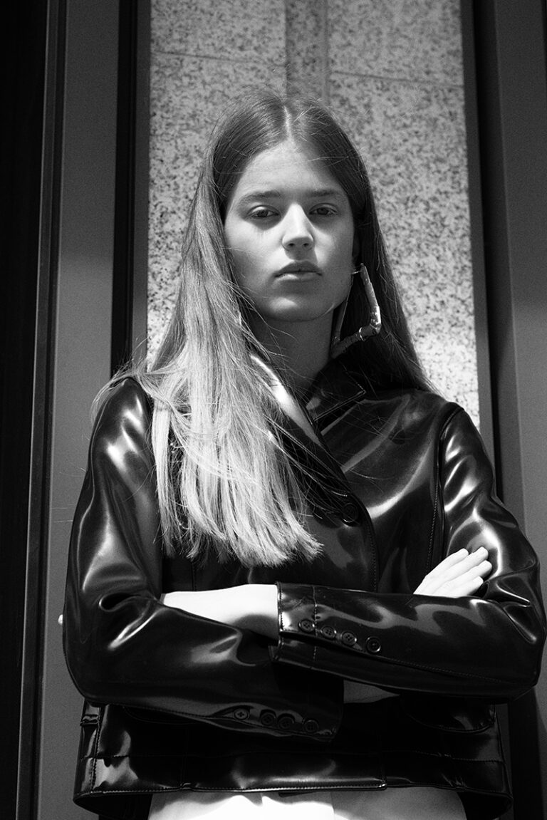 Modella Juta in bianco e nero durante un test per Supreme Management Milano, ritratta a tre quarti con giacca in pelle lucida e braccia conserte davanti a una parete di pietra, fotografata da Marius Cruceriu.