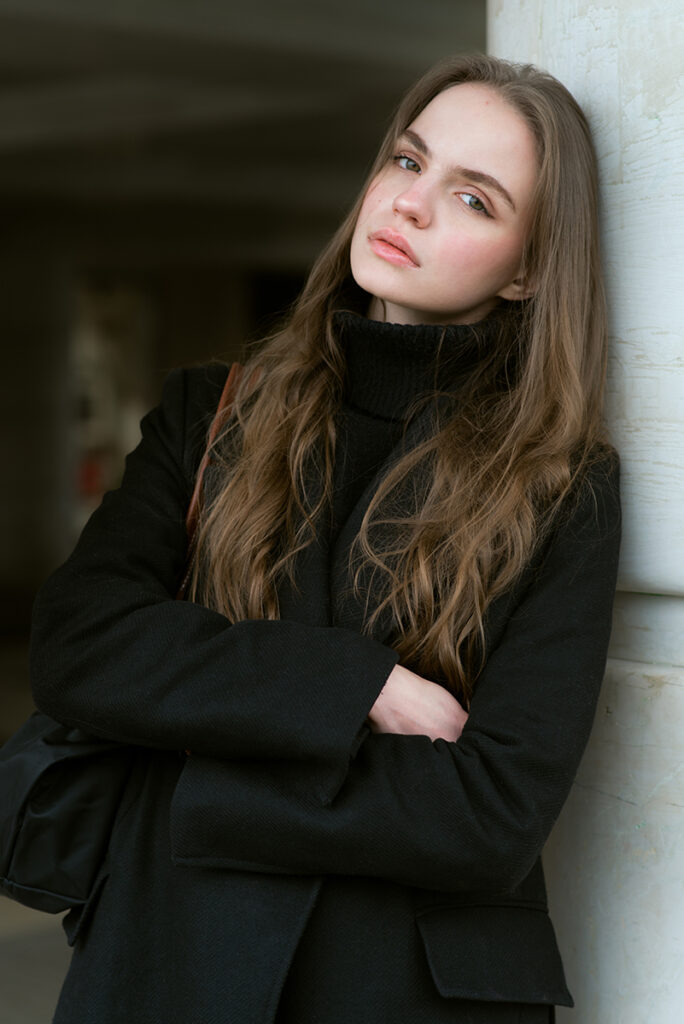 Modella Liddy per Novastars Models, ritratto close-up a colori con cappotto nero e dolcevita, posa appoggiata a una colonna, sguardo rilassato, scatto di Marius Cruceriu.