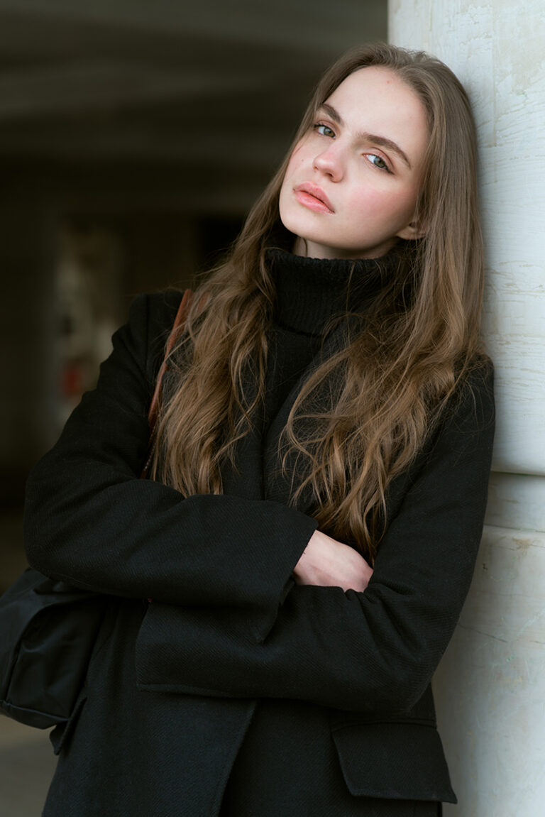 Modella Liddy per Novastars Models, ritratto close-up a colori con cappotto nero e dolcevita, posa appoggiata a una colonna, sguardo rilassato, scatto di Marius Cruceriu.