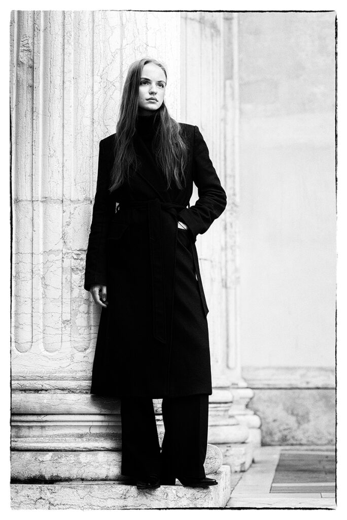 Modella Liddy per Novastars Models, scatto in bianco e nero a figura intera appoggiata a una colonna, con cappotto nero lungo ed espressione intensa, fotografata da Marius Cruceriu.