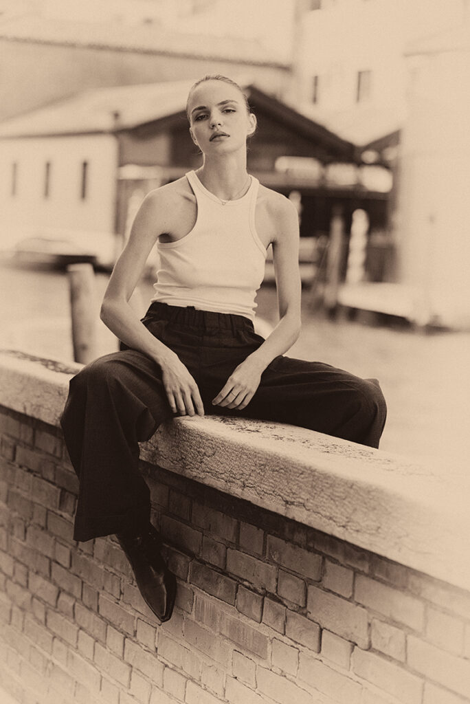 Modella Lidyia per Novastars Models a Venezia, seduta sul bordo di un canale in posa rilassata con canotta bianca e pantaloni scuri, fotografata da Marius Cruceriu in bianco e nero seppia.