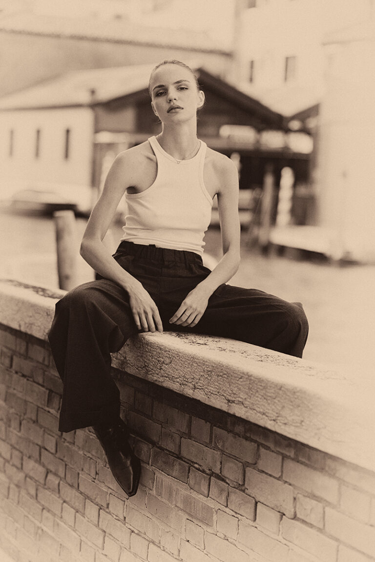 Modella Lidyia per Novastars Models a Venezia, seduta sul bordo di un canale in posa rilassata con canotta bianca e pantaloni scuri, fotografata da Marius Cruceriu in bianco e nero seppia.