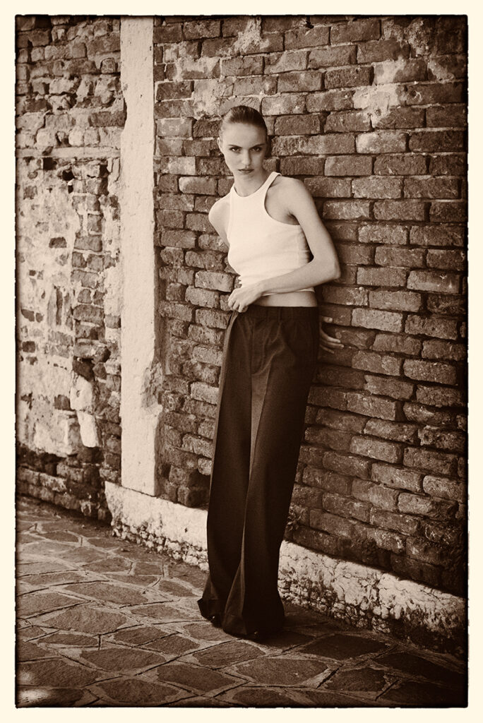 Modella Lidyia per Novastars Models a Venezia, ritratta a figura intera in bianco e nero appoggiata a un muro di mattoni, con pantaloni ampi scuri e top bianco smanicato, fotografata da Marius Cruceriu.