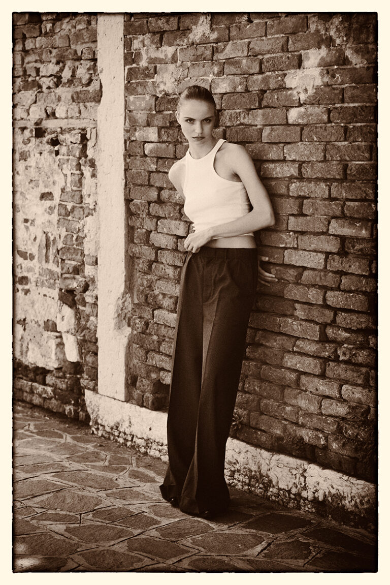 Modella Lidyia per Novastars Models a Venezia, ritratta a figura intera in bianco e nero appoggiata a un muro di mattoni, con pantaloni ampi scuri e top bianco smanicato, fotografata da Marius Cruceriu.