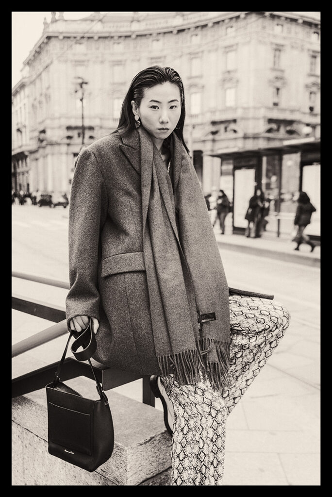 Modella Lu a Milano per Row Model Management, ritratto street style in bianco e nero, posa seduta in Piazza Cordusio con cappotto oversize e borsa, fotografata da Marius Cruceriu.
