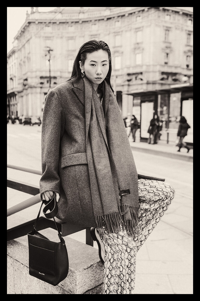 Modella Lu a Milano per Row Model Management, ritratto street style in bianco e nero, posa seduta in Piazza Cordusio con cappotto oversize e borsa, fotografata da Marius Cruceriu.