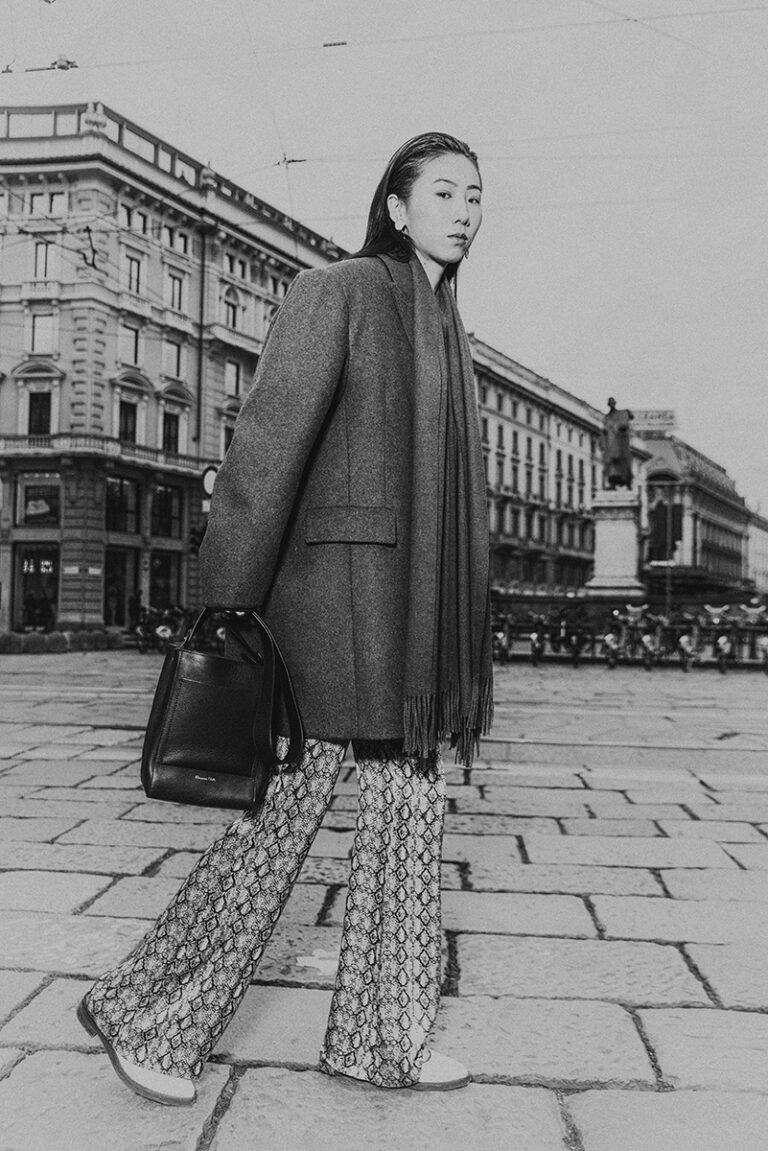 Modella Lu a Milano per Row Model Management, scatto street style in bianco e nero a figura intera in Piazza Cordusio, look con cappotto e pantaloni pitonati, fotografata da Marius Cruceriu.