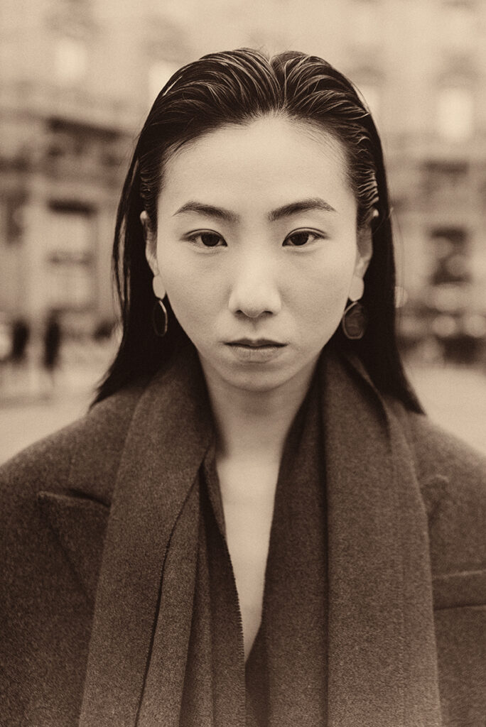 Modella Lu a Milano per Row Model Management, ritratto close-up in bianco e nero con sguardo diretto e cappotto scuro, test model realizzato da Marius Cruceriu.