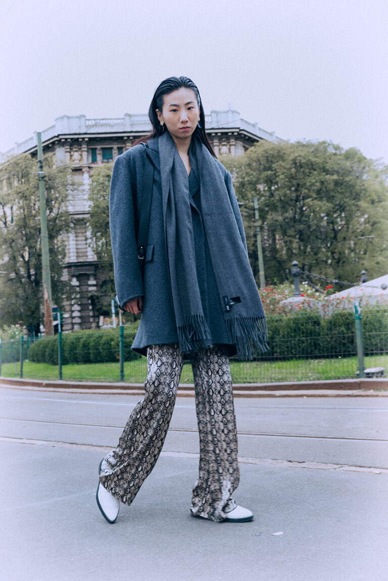 Modella Lu a Milano per Row Model Management, ritratto street style a colori a figura intera in Piazza Cordusio, look con cappotto grigio oversize, sciarpa e pantaloni pitonati, fotografata da Marius Cruceriu.