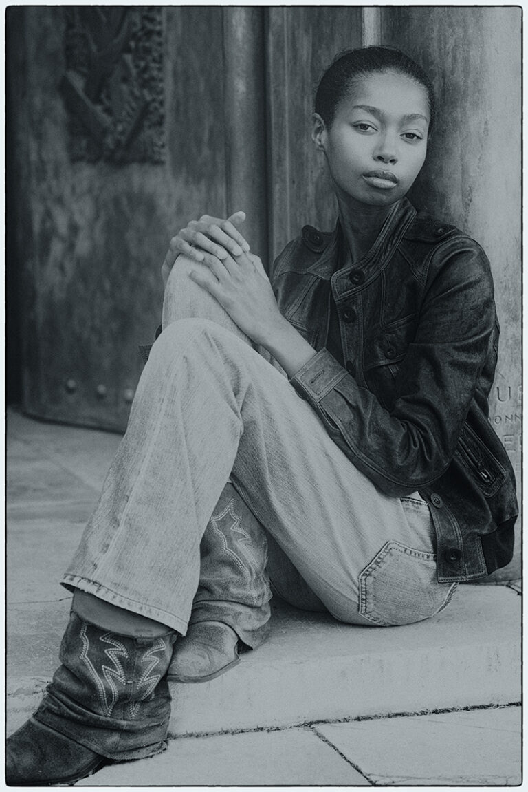 Modella Zoe a Parigi per The Face Paris, ritratto a figura intera seduta in esterno, bianco e nero, indossa giacca in pelle marrone e jeans con stivali, sguardo intenso, test model realizzato da Marius Cruceriu.