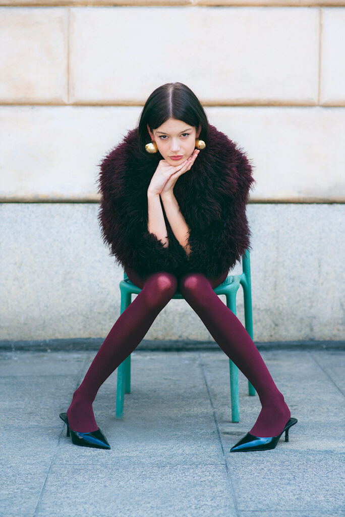 Modella Zuzanna durante un test per Supreme Management Milano, seduta su una sedia verde con posa a gambe divaricate, collant bordeaux, cappotto in pelliccia scuro e décolleté nere a punta, fotografata da Marius Cruceriu.