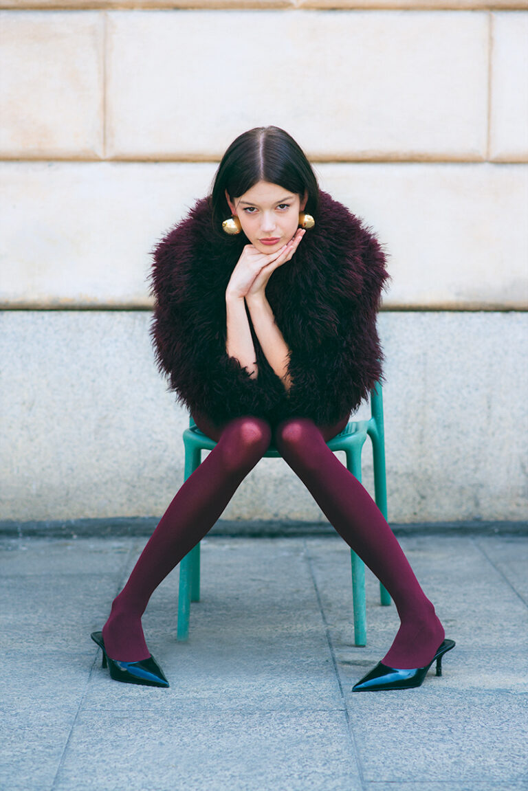 Modella Zuzanna durante un test per Supreme Management Milano, seduta su una sedia verde con posa a gambe divaricate, collant bordeaux, cappotto in pelliccia scuro e décolleté nere a punta, fotografata da Marius Cruceriu.
