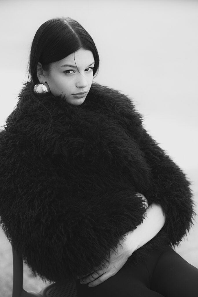 Primo piano della modella Zuzanna avvolta in un cappotto nero in pelliccia, seduta e con le mani intrecciate sul ventre durante un test per Supreme Management Milano, fotografata da Marius Cruceriu in bianco e nero.