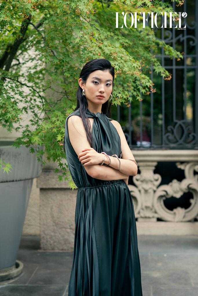 Modella asiatica in posa per l'editoriale "Of Shadows and Silk" su L'Officiel Baltics, indossa un abito verde scuro plissettato, braccia conserte e gioielli minimal, fotografata in esterno da Marius Cruceriu.