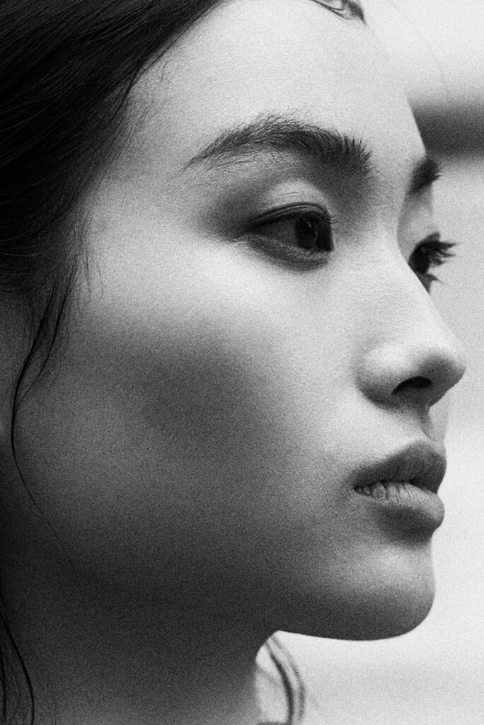 Primo piano (close-up) del viso della modella Liu in bianco e nero con grana evidente (stile pellicola Fuji Neopan 1600). Dettaglio su pelle, occhi e sopracciglia per l'editoriale "Of Shadows and Silk" di L'Officiel Baltic. Espressione intensa e introspettiva. Fotografia Marius Cruceriu.