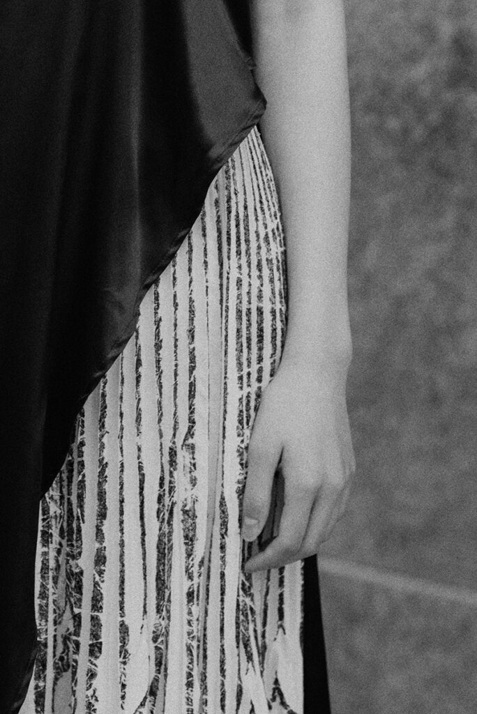Dettaglio in bianco e nero delle mani della modella Liu su tessuto plissettato e seta. Editoriale "Of Shadows and Silk" per L'Officiel Baltic. Focus su texture e contrasti di luce. Fotografia Marius Cruceriu.
