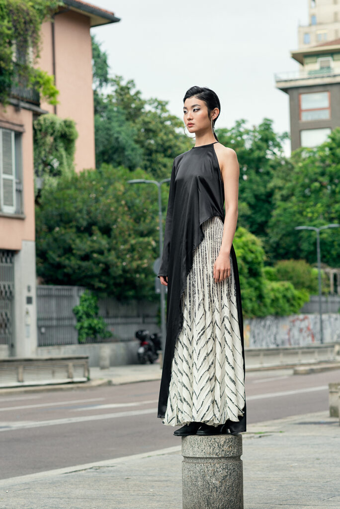 Modella Liu in posa statuaria su un basamento in pietra a Milano, scatto a figura intera per l'editoriale "Of Shadows and Silk" di L'Officiel Baltic. Indossa un look asimmetrico di Mine de Rien Couture. Fotografia di Marius Cruceriu.