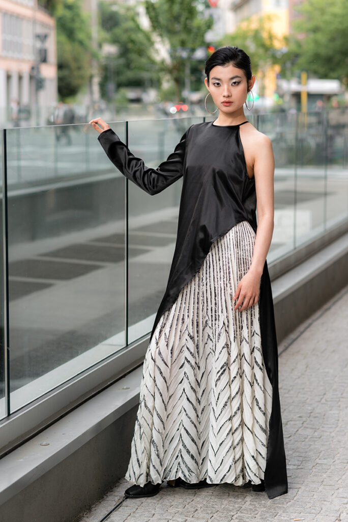 Ritratto a figura intera della modella Liu per L'Officiel Baltic, in posa accanto a una balaustra in vetro che riflette la città. Indossa un abito di Mine de Rien Couture con top in seta nera asimmetrico e gonna plissettata a stampa geometrica bianca e nera. Sfondo urbano moderno a Milano. Fotografia Marius Cruceriu.