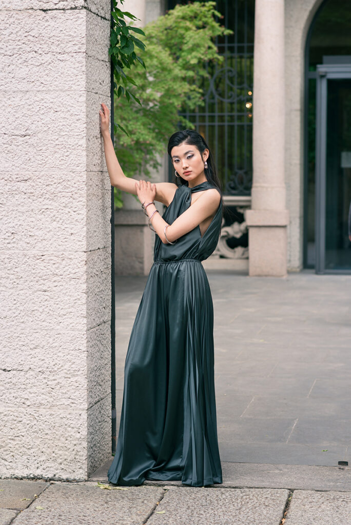 Ritratto moda a figura intera della modella Liu appoggiata a una colonna in pietra a Milano. Indossa una tuta verde scuro (jumpsuit) plissettata di Mine de Rien Couture con gioielli in argento. Editoriale per L'Officiel Baltic, fotografia Marius Cruceriu.