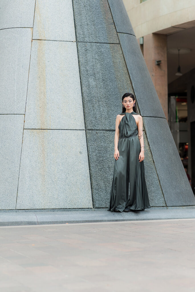Ritratto a figura intera della modella Liu davanti a una struttura architettonica moderna in pietra grigia. Indossa una tuta verde scuro (jumpsuit) con scollo all'americana di Mine de Rien Couture. Editoriale L'Officiel Baltic, fotografia Marius Cruceriu.
