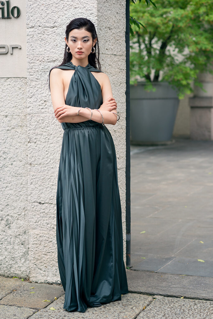 Modella Liu in posa statuaria a braccia incrociate davanti a un muro in pietra bugnata a Milano. Indossa una jumpsuit verde scuro plissettata di Mine de Rien Couture con gioielli minimal in argento. Editoriale L'Officiel Baltic, fotografia Marius Cruceriu.