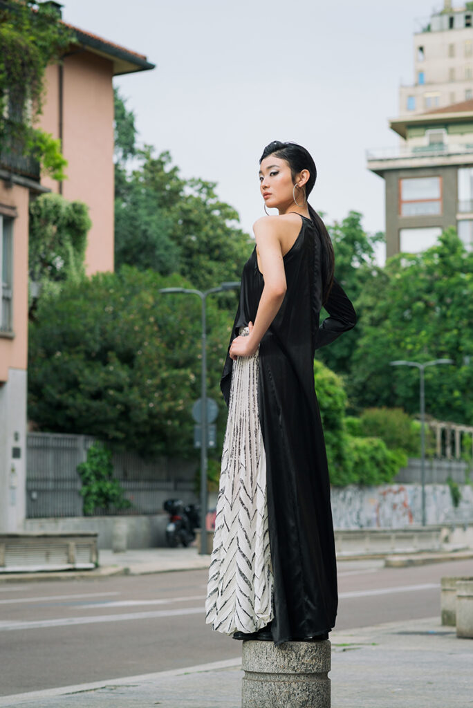 Ritratto a figura intera della modella Liu in strada a Milano per L'Officiel Baltic. Posa di schiena con torsione verso la camera, indossa un outfit Mine de Rien Couture con top nero lungo e gonna bianca plissettata a motivo geometrico. Sfondo urbano con edifici e alberi. Fotografia Marius Cruceriu.