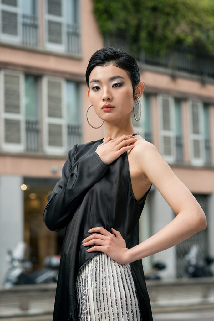 Ritratto a mezza figura della modella Liu per L'Officiel Baltic, editorial "Of Shadows and Silk". Posa "power pose" con mano sul fianco, orecchini a cerchio grandi e makeup glitterato. Styling urbano chic. Fotografia Marius Cruceriu.