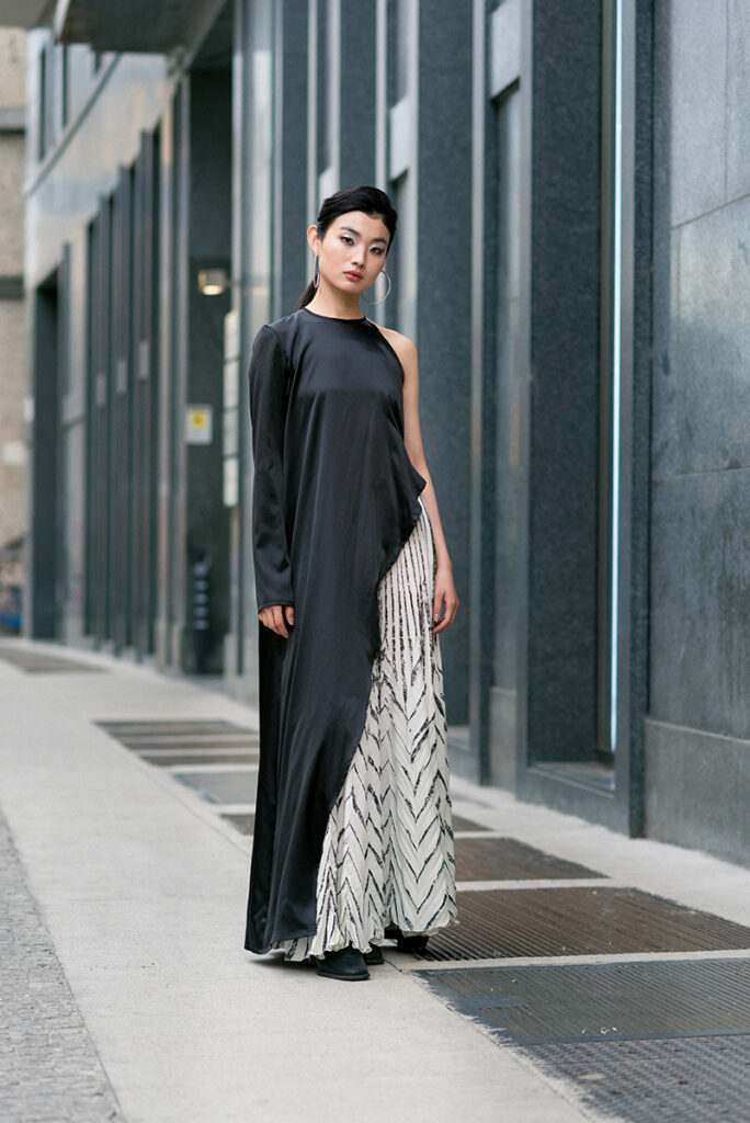 Ritratto a figura intera della modella Liu in un contesto urbano moderno a Milano per l'editoriale "Of Shadows and Silk" su L'Officiel Baltic. Indossa un abito asimmetrico nero e bianco di Mine de Rien Couture. Sfondo con colonne e architettura contemporanea. Fotografia Marius Cruceriu.