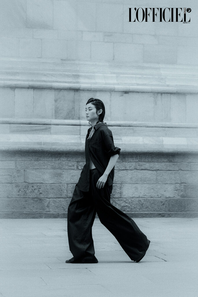 Pagina pubblicata su L'Officiel Baltic dell'editoriale "Of Shadows and Silk". Scatto a figura intera in bianco e nero della modella Liu che cammina indossando pantaloni ampi e camicia nera. Fotografia e art direction di Marius Cruceriu.
