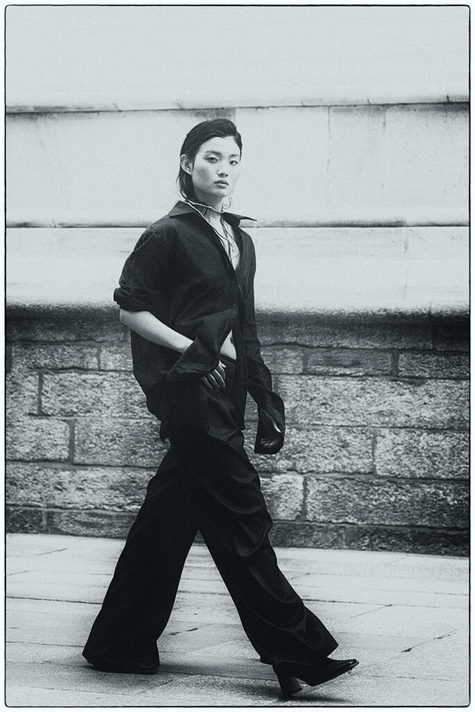 Modella Liu per l'editoriale "Of Shadows and Silk" pubblicato su L'Officiel Baltic, scatto a figura intera in bianco e nero mentre cammina con look maschile oversize, fotografia e direzione artistica di Marius Cruceriu.